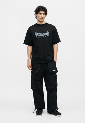 Joven con pelo rizado que lleva una camiseta negra con gráfico y pantalones cargo negros, de pie sobre un fondo blanco.