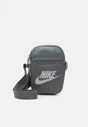 Petit sac à épaule Nike gris avec poche avant en filet, logo blanc, fermeture éclair et bandoulière réglable sur fond uni.