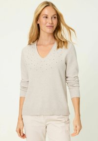 Hellbeiger V-Ausschnitt-Pullover aus weichem Material, mit einem gerippten Saum und Bündchen, verziert mit silbernen Nieten rund um den Ausschnitt.