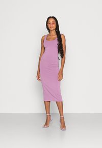 Robe midi sans manches de couleur violette au design ajusté, fabriquée en tissu extensible. Le modèle porte des sandales à talons transparentes, se tenant sur un fond uni.