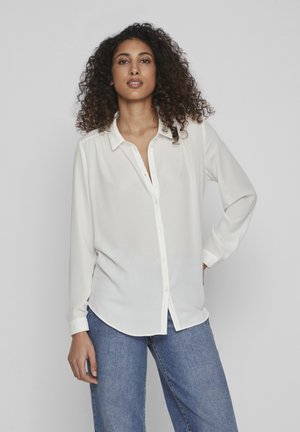 Blusa blanca de manga larga con botones en la parte delantera y cuello, hecha de un tejido ligero. Se lleva con jeans de denim azules, que tienen un corte relajado.