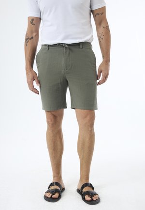 Mand iført olivengrønne shorts, hvid t-shirt, sorte sandaler og synlige tatoveringer på arme og ankel.