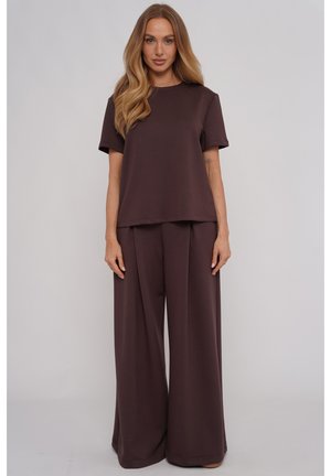 Kobieta z długimi blond włosami, ubrana w luźny ciemnobrązowy krótki top i szerokie spodnie, stojąca na tle jednolitego jasnego tła.