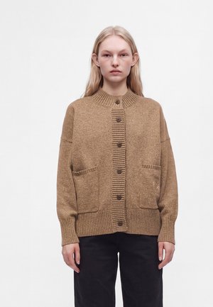 Jeune femme aux cheveux clairs porte un cardigan marron boutonné avec deux poches avant et un pantalon noir sur un fond blanc uni.