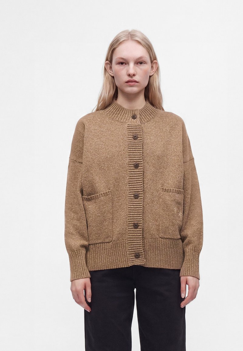 Jeune femme aux cheveux clairs porte un cardigan marron boutonné avec deux poches avant et un pantalon noir sur un fond blanc uni.