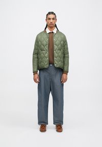 Jachetă verde matlasată cu nasturi prin presare, pulover maro dedesubt, blugi largi albaștri și pantofi din piele maro. Include o cămașă cu guler.