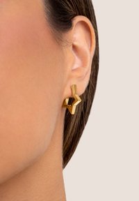 Nahaufnahme eines Ohrs einer Person, die einen goldenen, sternförmigen Ohrring vor neutralem Hintergrund trägt.