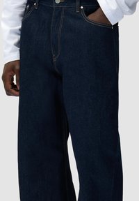 Jeans in denim blu scuro con cuciture visibili, indossati da una persona con una mano in tasca e l'altra lungo il corpo.