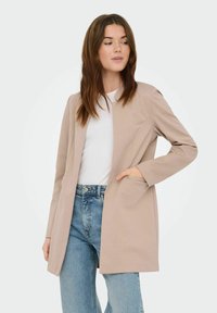 Blazer long beige au design minimaliste, présentant une seule ouverture avant, des poches latérales et une texture de tissu lisse, porté sur une chemise blanche.
