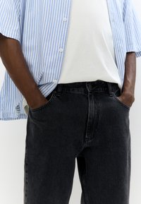 Camicia button-up con righe blu chiaro e bianche, abbinata a jeans neri in denim. Il tessuto sembra testurizzato e la vestibilità è rilassata.