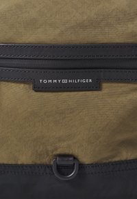 Olivgrön tygryggsäck med en svart zip-ficka, som har en svart läderetikett med "TOMMY HILFIGER" präglat. Metallbeslag som accentuerar.