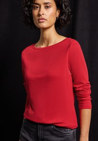 Rotes Langarmoberteil aus glattem Stoff mit Rundhalsausschnitt. Die Ärmel sind am Handgelenk leicht gerafft. Kombiniert mit dunkelgrauen Jeans.