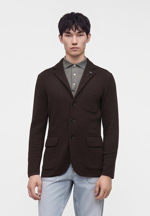 Mann trägt ein dunkelbraunes, geknöpftes Sakko über einem grauen Polo mit hellblauen Jeans, steht vor einem schlichten weißen Hintergrund.