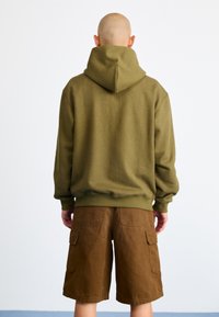 Sweatshirt verde-azeitona com um capuz grande e punhos canelados, combinado com calções cargo castanhos que apresentam vários bolsos. Materiais texturizados evidentes.