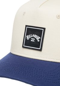 Beige pet met een marineblauwe klep, voorzien van een zwart vierkant patch met witte tekst en logo. De stof heeft een gestructureerd patroon.