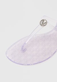 Gennemsigtig lavendel flip-flop med et tekstureret cirkulært mønster på sålen og en transparent rem med et sølvfarvet logoaccent.