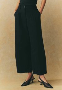 Pantaloni neri a gamba larga con vita alta, pieghe e tasche frontali. Abbinati a décolleté slingback in rete nera con punta affusolata.