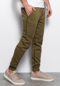 Olivgrüne Sweatpants mit schwarzen Nähten, schmaler Schnitt, Seitentaschen und elastischem Bund, kombiniert mit hellgrauen Sneakers.