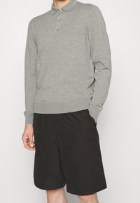 Polo gris en tricot avec un col, trois boutons et un logo sur la poitrine. Associé à un short noir arrivant aux genoux avec une ceinture élastique.