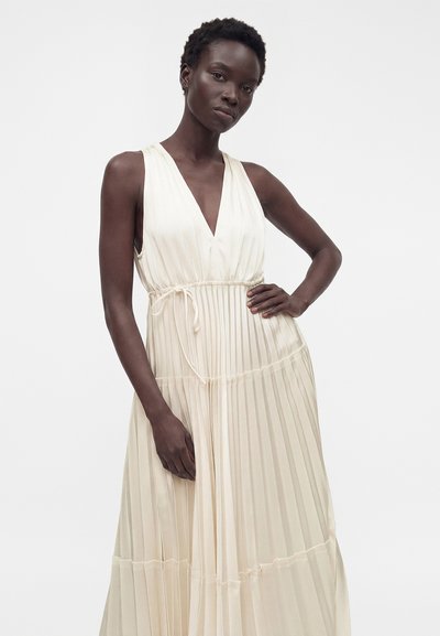 Femme aux cheveux courts portant une robe longue plissée crème sans manches avec décolleté en V et taille nouée, posant devant un fond clair uni.