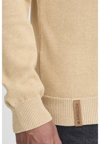 Suéter de punto beige con puños y dobladillo acanalados. La etiqueta dice "JEANS CODE". La textura parece suave con sutiles motas en toda su superficie.