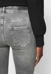 Jeans cinzentos desbotados com corte slim, apresentando um bolso traseiro e uma etiqueta preta com o logótipo "ONLY" na cintura. Material de denim texturizado.