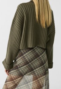 Olijfgroen geribbeld cropped truitje met losse mouwen, gelaagd over een doorschijnende geruite rok in aardetinten en subtiele patronen.