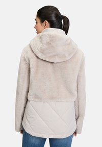 Veste beige clair avec une partie supérieure en peluche et une partie inférieure matelassée. Elle possède une capuche et des manches longues.