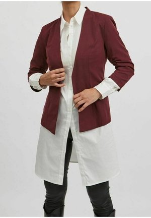 Vrouw in een bordeauxrode blazer over een lange witte blouse met knopen, zwarte broek en laarzen, die de voorkant van de blazer aanpast.