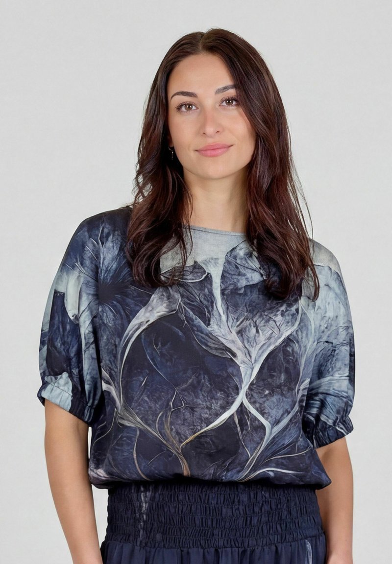 Femme aux longs cheveux bruns portant un chemisier ample bleu foncé avec de grands motifs floraux abstraits et une jupe assortie sur un fond uni.