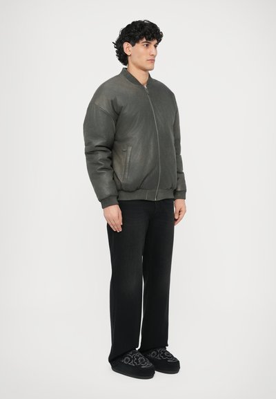 Tamsiai alyvuogių žalios spalvos bombonešio stiliaus striukė su priekine užtrauktuku, ribotos apyrankėmis ir apačia, dėvima su juodomis džinsais ir raštuotais juodais šlepetėmis.