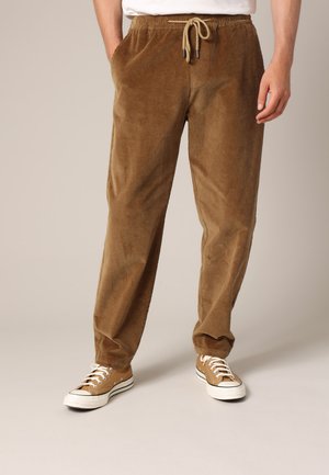 Persoon draagt bruine corduroy broek met elastische taille en trekkoord, gecombineerd met beige sneakers met witte zolen en veters.