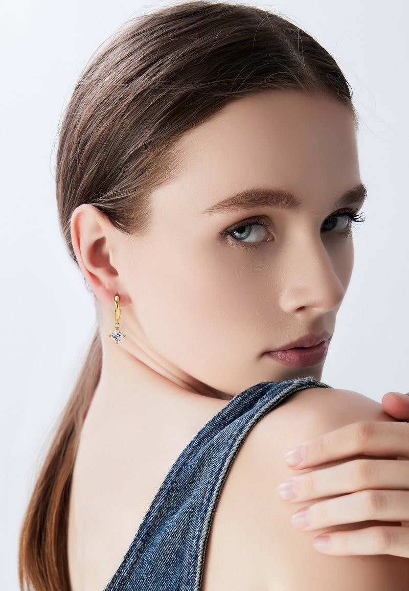 Zlatna bešavna naušnica s prozirnim kamenim privjeskom, nosi je model s srednje smeškom kosom, mekom kožom i traper košuljom.