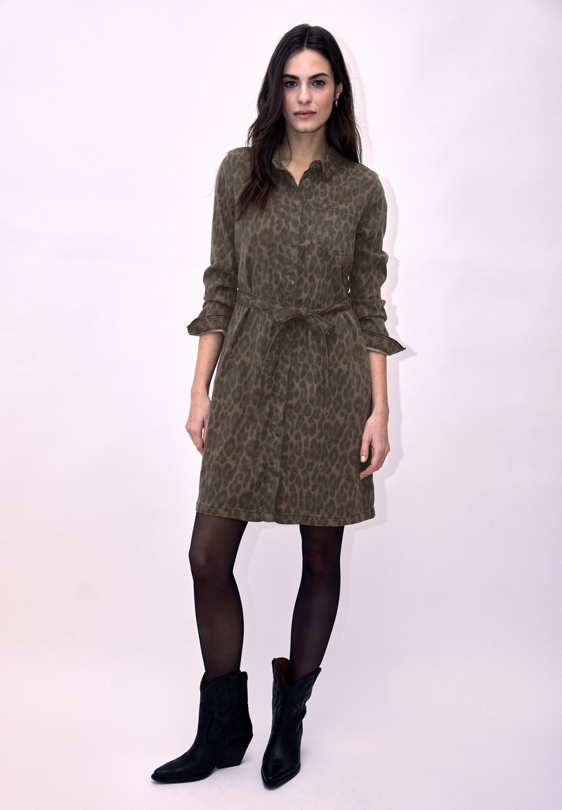 Femme portant une robe boutonnée imprimé léopard marron avec une ceinture à la taille, des collants noirs et des bottines noires, debout devant un fond clair.