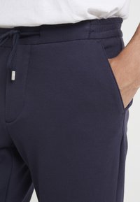 Marineblå joggingbukser lavet af et struktureret stof, med elastisk talje og snor. Har sidelommer og syninger langs sømmene.