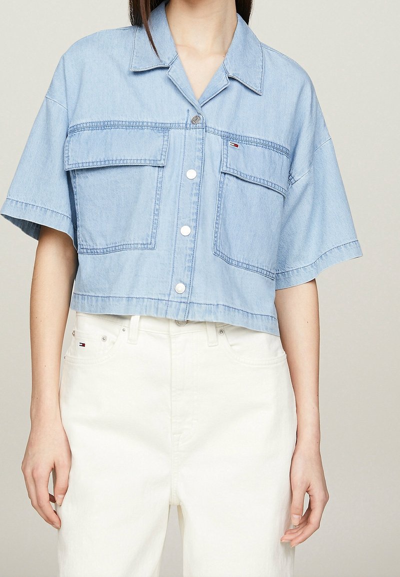 Light blue cropped denim shirt with a collar, short sleeves, two front pockets, and button closure, featuring a small logo accent.