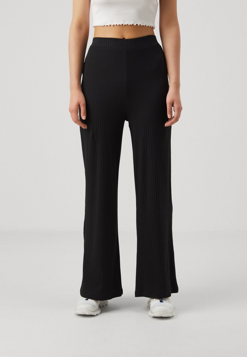 Femme portant un pantalon large côtelé noir taille haute et des baskets blanches, debout devant un fond clair uni.