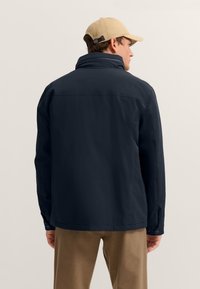 Marineblaue wasserfeste Jacke mit hohem Kragen, geknöpften Manschetten und geradem Schnitt. Getragen über hellbrauner Hose, von hinten gezeigt.