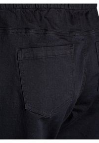 Pantalones de mezclilla negra con cintura elástica, una textura suave y dos bolsillos traseros con forma angular. Costuras a contraste visibles.