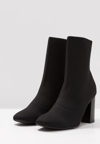 Bottes noires à la cheville en textile tricoté ajusté, avec un bout pointu et un design à talon carré, présentant une texture lisse.