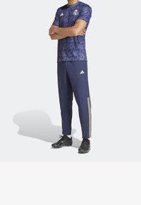 Sportovní oblečení Adidas s vzorovaným fialovým tričkem a námořnickými kalhotami se zlatými pruhy, doplněné černými fotbalovými kopačkami.