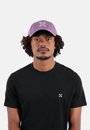 Homme aux cheveux bouclés portant une casquette lilas et un t-shirt noir avec un petit logo blanc sur la poitrine, faisant face à l'avant sur un fond uni.