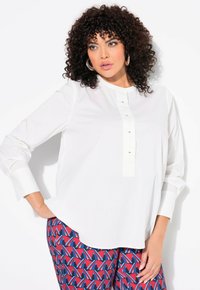 Chemise blanche à col rond, manches longues, avec une patte de boutonnage comportant de petits boutons noirs, accompagnée d'un pantalon à motifs rouges et bleus.