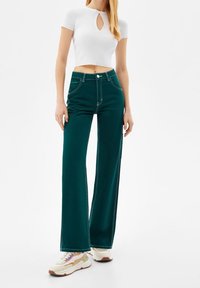 Nainen, joka on pukeutunut valkoiseen lyhythihaiseen cropped-paitaan ja korkeavyötäröisiin tummanvihreisiin leveisiin housuihin, valkoiset lenkkarit jalassa valkoista taustaa vasten.