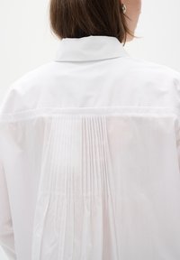 Bílá košile s přehozeným límečkem a vertikálními sklady na zadní straně, vyrobená z hladké látky, s čistým a minimalistickým designem.