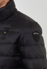 Giubbotto trapuntato nero con colletto alto, etichetta del logo giallo Blauer USA sul petto e patch nera Blauer sulla manica superiore.
