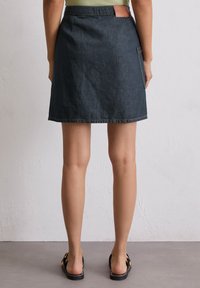 Jupe en jean A-line bleu foncé avec un léger éclat. Présente une poche arrière et une étiquette en cuir à la taille. L'ourlet se termine par des coutures discrètes.