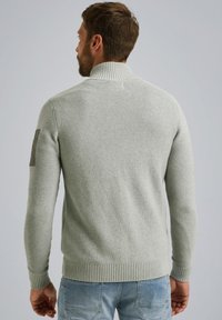 Lichtgrijze gebreide trui met een hoge kraag, geribbelde manchetten en zoom. Bevat een gestructureerd patroon en een patch op de linker schouder.