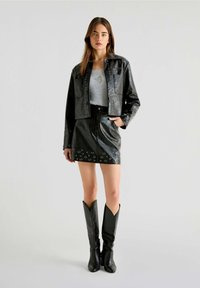 Chaqueta corta de piel sintética negra con detalles de ojales, combinada con una falda a juego. Las botas presentan un diseño puntiagudo en la parte superior.