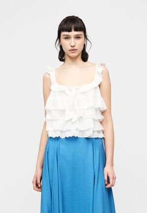 Νεαρή γυναίκα φορεί λευκό αμάνικο τοπ με βολάν και μπλε υφασμάτινη φούστα, στέκεται μπροστά από ένα απλό φωτεινό φόντο.